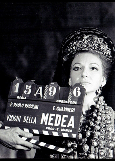 A black and white photograph of a seated Maria Callas, in the role of Medea, with a clapperboard shutting in front of her. 