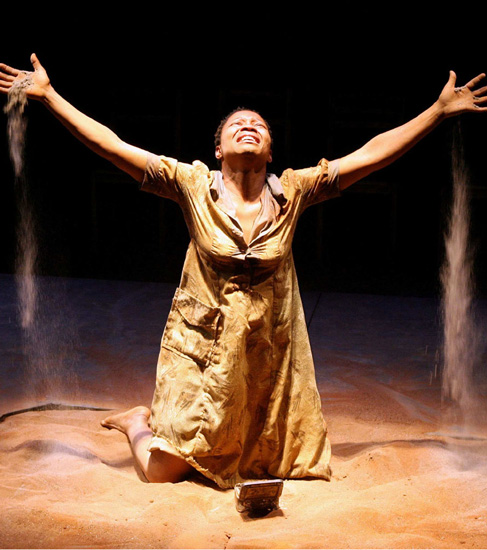 Actor Jabulile Tshabalala as Elektra - kneeling in sand with head and arms raised, sand falling from her fingers