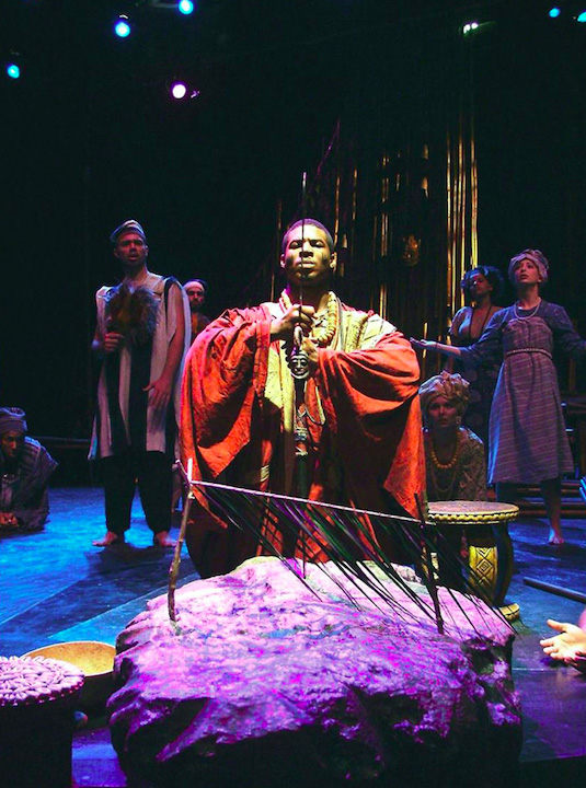 A man kneels centre stage, wearing red ceremonial robes, holding a sword in front of his face, his eyes closed: production photograph of the 2004 performance of The Gods Are Not To Blame; Ohio University's Forum Theatre 