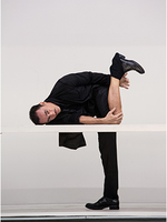 Photograph of a male dancer, dressed all in black against a blank white background; he is stood on one leg with his body and face lying on a white table. He is looking to camera; his other leg is bent upwards and is bare from knee to ankle.