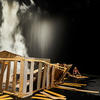 photograph of Phia Ménard’s play Mother House depicting a broken cardboard structure in the foreground, with smoke or dust falling from above the structure, and a figure seated on the floor in the background