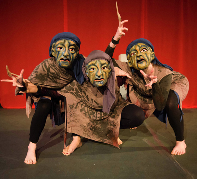 Three actors in theatrical masks, performing as Furies, crouching as a group in front of a red curtain