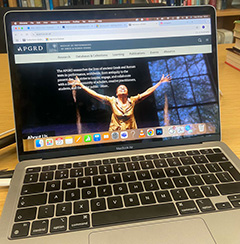 Photograph of a laptop on a desk, with bookshelves in the background. The laptop displays the homepage of the new APGRD website