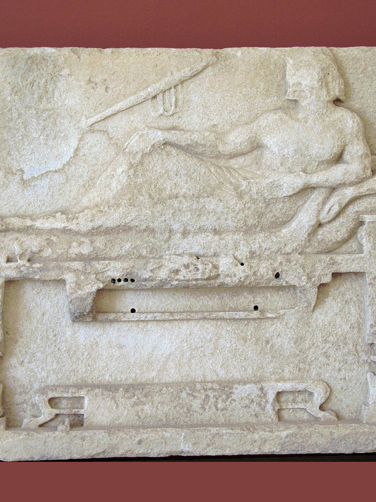 Photograph of an ancient Greek marble relief depicting a man reclining on a bed and possibly some musical instruments